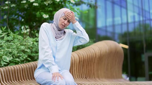 Young muslim female in hijab is suffering from a headache while sitting on a bench on the street