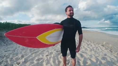 Surfer Stands on Beach Holding Red Surfboard