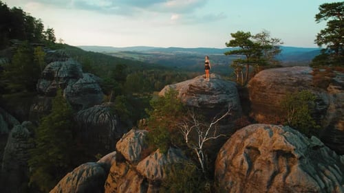 A lone hiker stands atop a rock formation at sunset, surrounded by towering cliffs and lush greenery