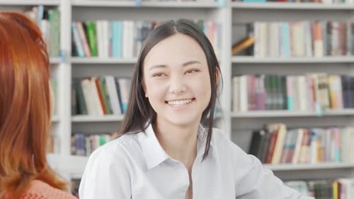 Fröhliche junge Asiatin lacht, während sie mit ihrer Freundin in einer lebendigen Bibliothek lernt