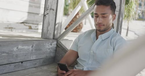 Thoughtful biracial man using smartphone sitting on porch of wooden beach house, in slow motion