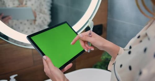 Primer plano de las manos femeninas usando una tableta negra con pantalla verde en el baño de la casa. Vista desde la parte trasera