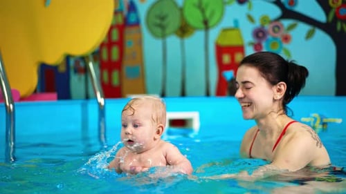 Adorable chubby baby boy making splashes in the pool. Woman is holding a child teaching him to swim.