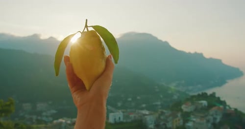 Close-up of a hand holding a ripe lemon with a sunlit backdrop of the Amalfi Coast, conveying the fr