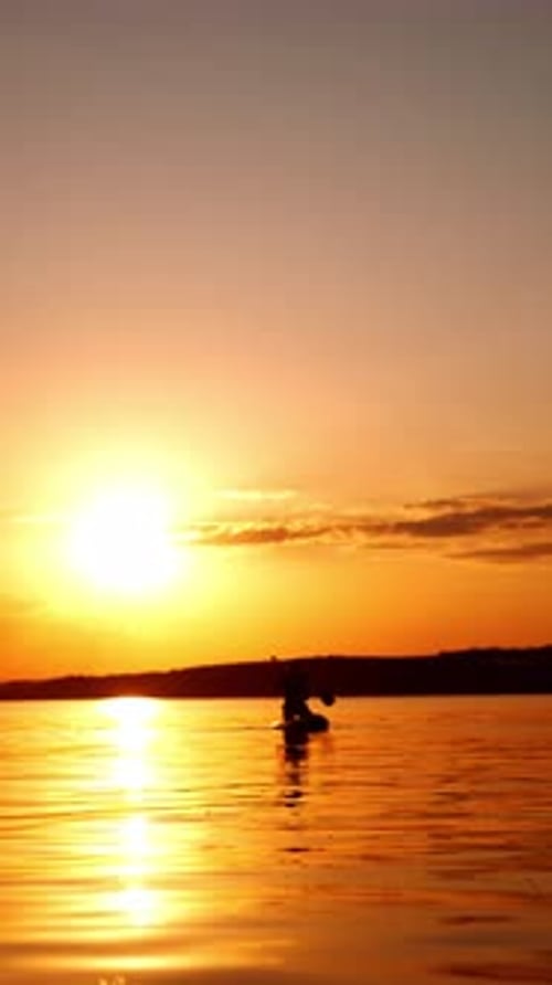 Person sits on the paddle board approaching camera. Sunset on the river for practicing sport.