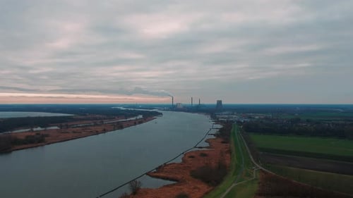 Drohnen-Hyperlapse eines Kraftwerks am Flussufer, umgeben von landwirtschaftlichen Feldern.