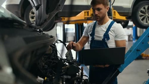 Handsome mechanic in uniform is using a laptop while repairing car in auto service