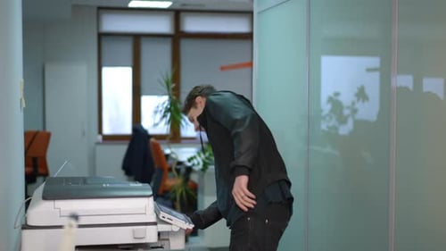 Junger Mann überprüft kaputten Drucker im Büro und denkt nach