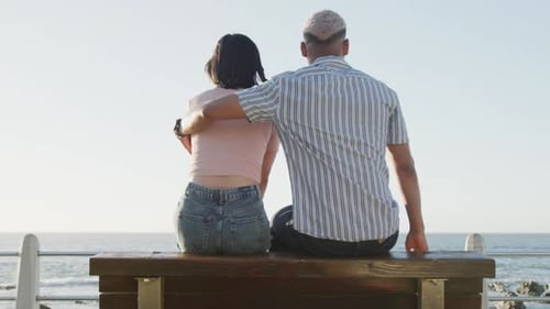 Happy biracial couple sitting on bench and embracing on promenade, in slow motion