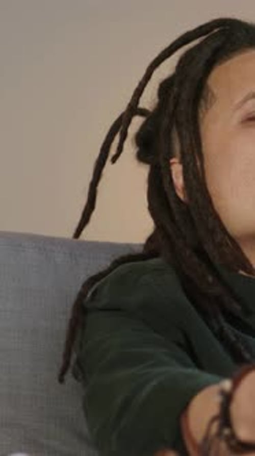 Vertical video: Sipping drink, man with dreads in green shirt pressing remote, adjusting TV at home