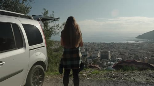 Car Journey, a woman admires the city and the sea from a height while standing near her car.