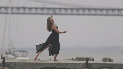 An Adult Woman in Black Dress Dancing on the Pier with a Bridge on the Background