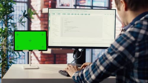 Man in Home Office Looking at Green Screen Tablet and Computer Typing Code