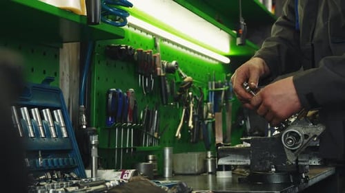 Auto mechanic repairs auto part steering rack in a large workshop filled with various tools hanging