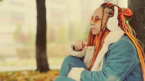 Young Beautiful Woman Listening to Music in Wireless Headphones Sitting Near Tree in City Park