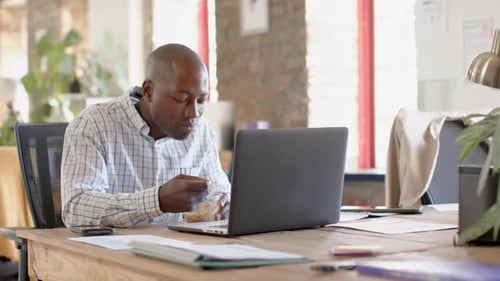 Happy african american casual businessman using smartphone and having lunch in office, slow motion