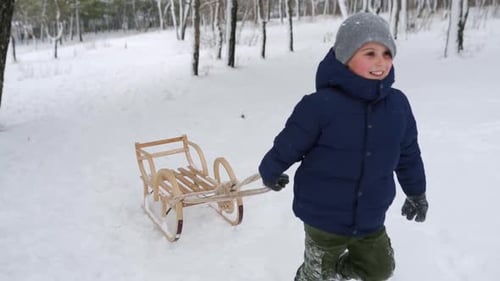 Süßer Junge in warmer Kleidung, der am verschneiten Wintertag Holzschlitten zieht Kleiner Kerl rodelt im Park