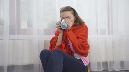 The woman drinking coffee leaning against the heater is cold.