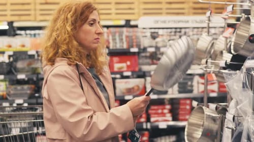 Red-haired woman in large supermarket chooses cast-iron frying pan in department of cooking utensils