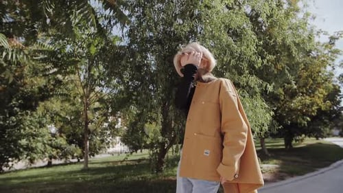 A Young Blonde Woman in a Yellow Trendy Coat Walks Down the Street