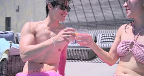 Couple sitting on towel poolside in swimwear, man raising citrus glass during talk to toast
