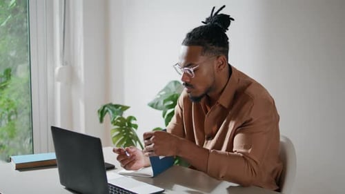Man Writes in Notebook at Desk with Laptop