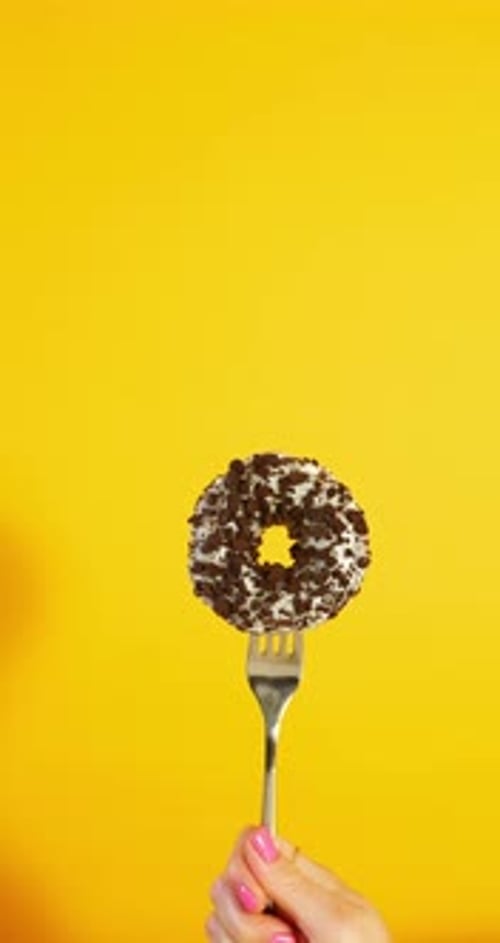 A hand with pink nails lifts a fork holding a white-glazed donut topped with cookie crumbles high ag