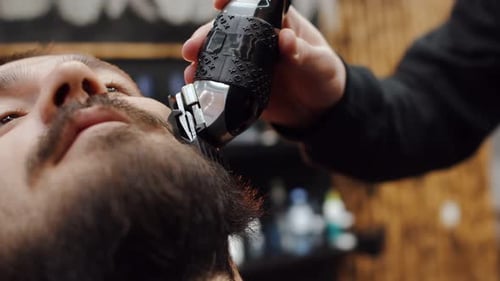Professional Barber Man in a Barbershop Cuts the Beard of a Young Hipster Client of Caucasian