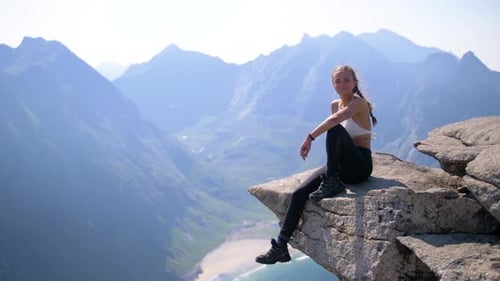 A pretty woman sits on a ledge high on a mountain overlooking the incredible scenery of Lofoten, Nor