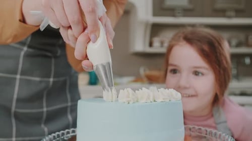 Mom and daughter decorate a freshly made layered cake with cream frosting. A curious girl smiles and