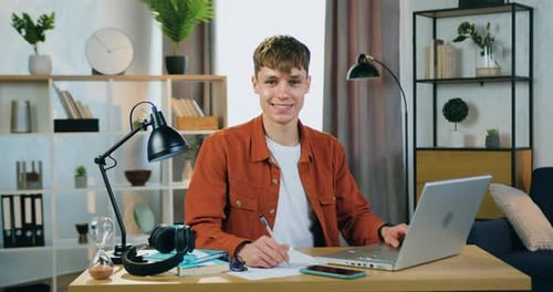 Portrait of young man which looking into camera when doing university home task sitting at his workp