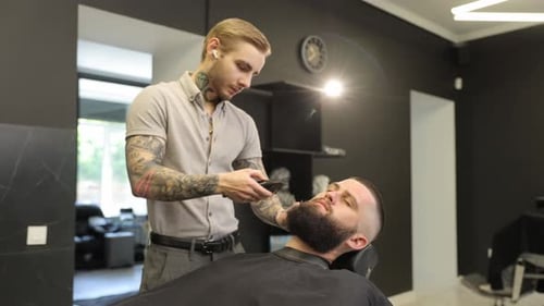 Tattooed Barber Trimming a Man's Beard with a Clipper