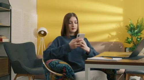 Serious Young Female Student Typing on Cell Phone Sitting at Table in Her Room