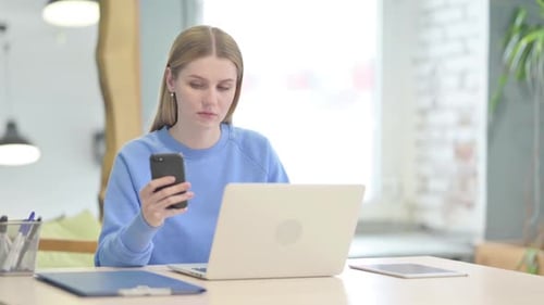 Young Woman Using Smartphone at Desk with Laptop