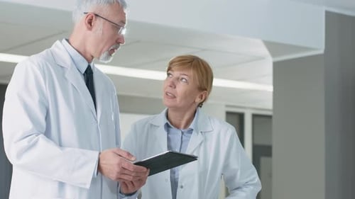 In the Hospital Doctors have Discussion while Using Tablet Computer. In the Background Patients and