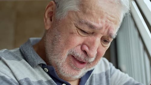 Close-Up Of Elderly Man With Gray Beard And Suffering Face Leaning Toward Iron Garage Bars, With