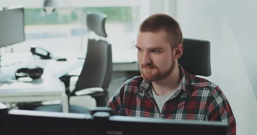 Ein junger bärtiger Mann sitzt an einem Computer, arbeitet im Büro, fühlt sich glücklich, Gesichtsmonitor online
