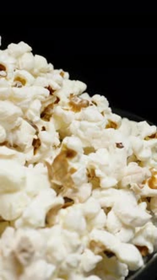Vertical video. A large plate with a heap of popcorn rotates against a black background. Close-up.