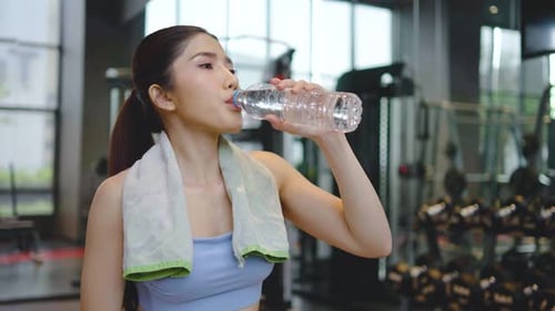 Portrait of tired asian woman in sportswear drinking water from bottle in gym.