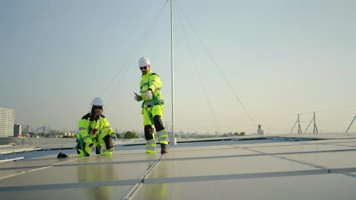 Two professional technicians inspect solar panels on a rooftop. They use digital tablets