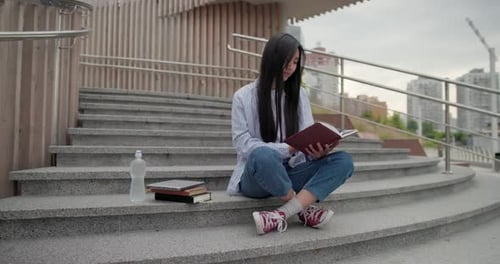 Education in University College Concept Young Lady Reading Notebook While Sitting Outdoors on Steps