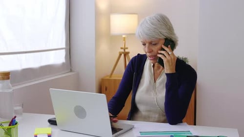 Mature Woman Using Laptop and Taking Notes While Talking on the Phone at Home Office