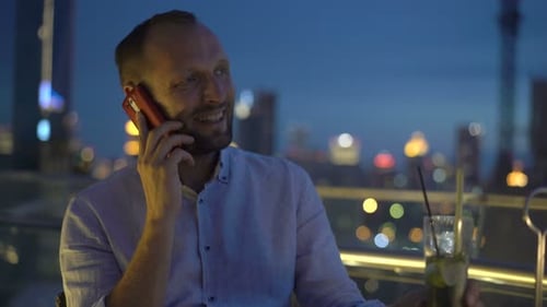 Young Man Talking on Cellphone and Drinking Cocktail on Terrace in Bar