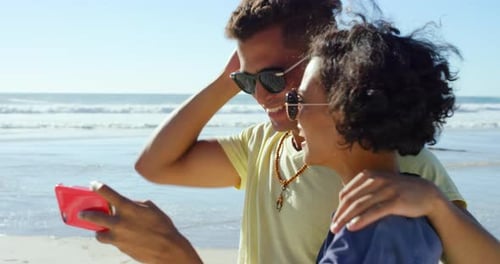 4k video footage of a young couple standing together on the beach and taking selfies on a cellphone
