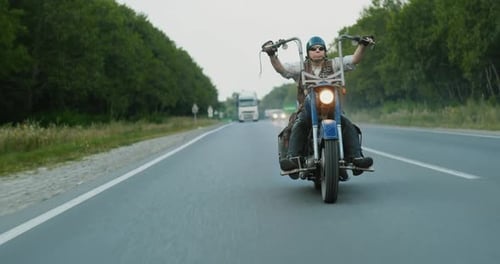 Lone Biker on a Custom Motorcycle Speeds Along an Asphalt Road That Runs Through the Forest in the