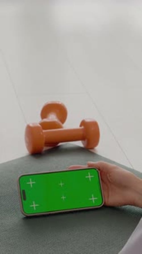 Vertical video Woman relaxes on gym mat using smartphone with green screen for workout