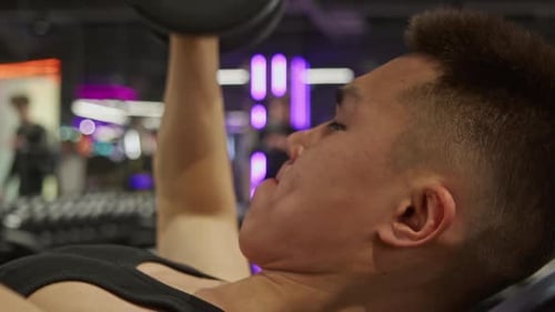 Ein junger asiatischer Mann trainiert im Fitnessstudio, liegt auf einer Bank und hebt dabei mehrere schwere Hanteln