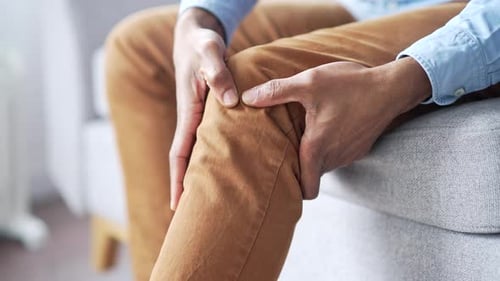 Close up. Male hands massaging knee leg muscles while sitting on sofa in living room at home. Senior