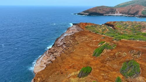 Orange bare rocks with some spots of greenery. Blue waterscape surrounds the rocky shore.