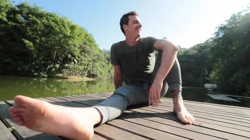 Happy man smiling and hanging outside in nature seated at deck next to lake